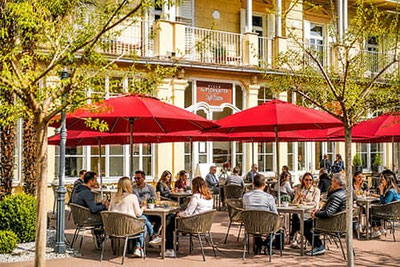 La terrazza soleggiata del Café Bistro Villa Aufschnaiter a Bolzano-Gries, con ombrelloni rossi e ospiti che si godono la colazione, l'aperitivo e momenti di piacere.
