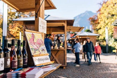 Colorate bancarelle al mercato contadino di Lagundo con prodotti regionali di qualità, bottiglie di vino e uno scenario autunnale: un punto d'incontro per i buongustai.