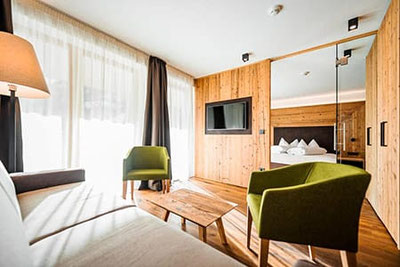 Heller Wohnbereich im Hotel Adlernest mit Holzwänden, gemütlichen Sesseln, Sofa, großem Fenster und Blick in das angrenzende Schlafzimmer.