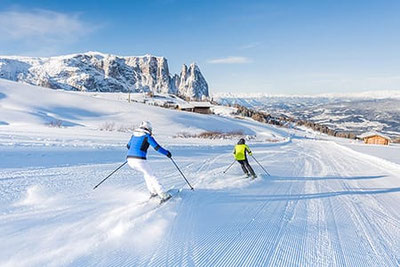 Sciatori su pista appena preparata sull’Alpe di Siusi davanti al massiccio dello Sciliar, giornata invernale soleggiata con vista sulle Dolomiti innevate.