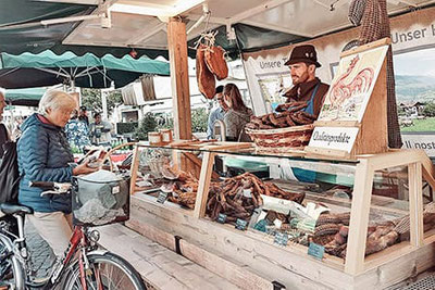 Bauernmarkt am Waltherplatz in Bozen: Ein Stand mit Südtiroler Speck, Würsten und regionalen Spezialitäten, freundlich bedient von Produzenten in Tracht.