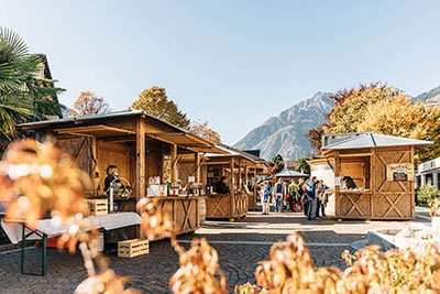 Bancarelle in legno del mercato dei sapori di Lagundo sullo sfondo autunnale delle montagne, i visitatori passeggiano tra i prodotti regionali e si godono la soleggiata atmosfera autunnale.