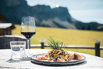 Piacere culinario durante le settimane dell'agnello della pecora dagli occhiali di Funes: pasta fatta in casa con ragù di agnello, un bicchiere di vino rosso e panorama sulle Dolomiti in un rifugio soleggiato.