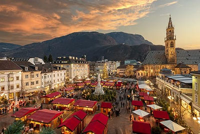 Il suggestivo mercatino di Natale di Bolzano al tramonto in Piazza Walther, con bancarelle illuminate a festa, albero di Natale e vista sul Duomo di Bolzano.