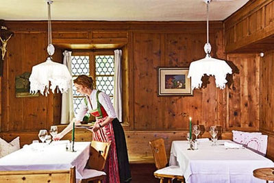 Maria, vestita con il tradizionale dirndl, apparecchia la tavola nell'accogliente sala in legno del ristorante Turmwirt a Gudon: atmosfera tradizionale, lume di candela e ospitalità altoatesina.
