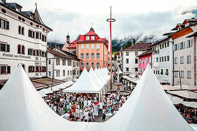 Der Marktplatz in Kaltern während der Veranstaltung „Kaltern ganz in Weiß“, mit festlich aufgebauten Zelten und einer großen Menschenmenge, die die besten Kalterer Weißweine verkostet und genießt.  
