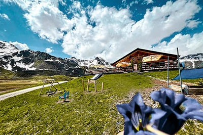 Lazaun Hütte auf dem Hochplateau im Schnalstal mit Sonnenterrasse, Spielplatz und Almwiesen, umgeben von alpiner Berglandschaft unter sommerlichem Himmel.
