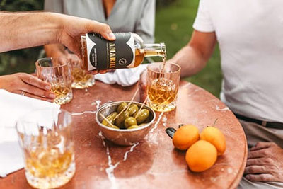 Amaro Amarill wird in Gläser eingeschenkt, dazu Oliven und frische Marillen auf einem Tisch – gesellige Genussmomente mit Südtiroler Marillen-Amaro im Freien