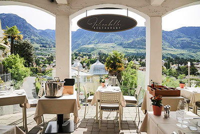 Terrazza del ristorante Felsenkeller con vista su Merano, tavoli apparecchiati con stile, palme e panorama montano: un posto in prima fila sulla Passeggiata Tappeiner.