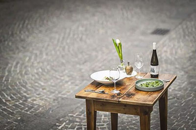 Tavolo in legno elegantemente apparecchiato all'aperto davanti al bistrot Speckeggele a Silandro, con vino, piatti freschi e decorazioni floreali per momenti di puro piacere.
