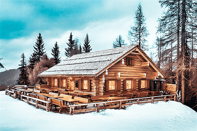 Verschneite Skihütte Ücia Picio Pré mit Sonnenterrasse und Holztischen, warm beleuchteten Fenstern und Blick auf die winterliche Berglandschaft am Kronplatz in Südtirol.