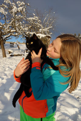 Blondes Mädchen im Skianzug schmiegt ihr Gesicht an eine schwarze Katze 
