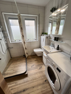 Modernes Badezimmer der Ferienwohnung „Auszeit Niedersfeld“ mit ebenerdiger Dusche, WC, Waschbecken und Waschmaschine. Große Fenster sorgen für Tageslicht, helle Fliesen und Holzoptikboden schaffen ein gepflegtes und freundliches Ambiente.