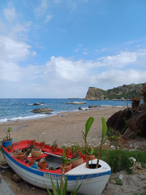 Auf dem Foto ist im Vordergrund ein kleines Boot, welches zur Dekoration genutzt wird mit Blumen zu sehen und im Hintergrund ist der Kiesstrand und das Meer zu sehen.
