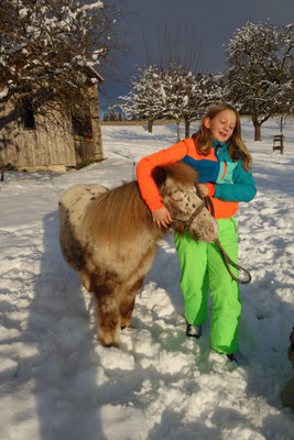 Ein blondes Mädchen umarmt den Hals eines weißen Ponys im verschneiten Garten am Biohof Stadler