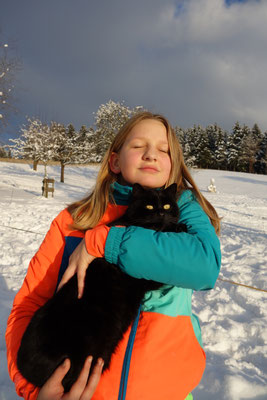 Ein Mädchen genießt mit geschlossenen Augen und einer schwarzen Katze im Arm die Wintersonne.
