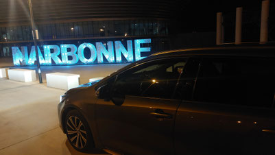 Véhicule de VTC RC VTC Narbonne stationné de nuit devant le panneau lumineux bleu Narbonne, service de transport privé Occitans, département de l'Aude.