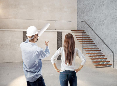 Bauleiter mit Helm und Bauplan zeigt auf eine freischwebende Treppe, Frau blickt in den offenen, modernen Raum