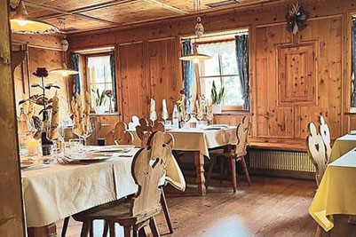 Accogliente sala rivestita in legno nell'osteria contadina Oberpartegger a Villandro con tavoli apparecchiati con cura, luci calde e vista sul paesaggio montano dell'Alto Adige.