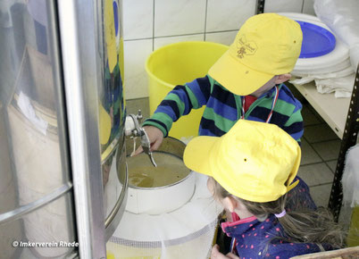 Zwei Kinder knieen vor einer Honigschleuder und sehen dem Honig zu