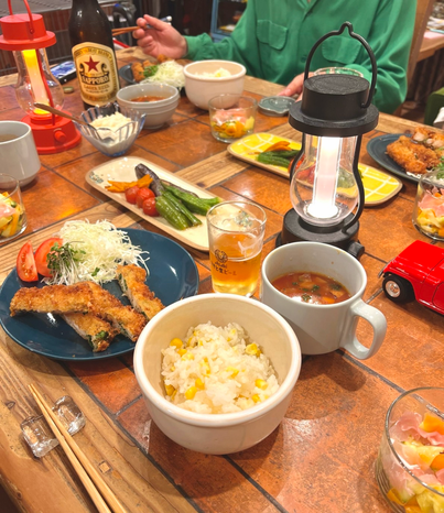 テーブルの上にはバルミューダの赤と黒のランタンが並び、お茶碗にはとうもろこしご飯、ブルーのお皿にはエビカツとキャベツの千切り、カットトマト、スープカップにはミネストローネ、そして夏野菜の揚げ浸しとビールなどが食卓を彩っています。