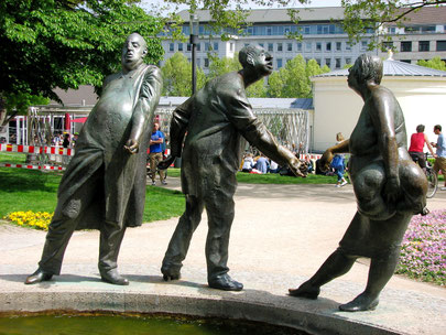 Sehenswürdigkeiten Aachen: Bronzefiguren stehen auf dem Brunnenrand. Manche der Figuren geben sich die Hand. Die Figuren zeigen zumeist Personen im mittleren Alter. Ihre Kleidung und Leibesfülle zeigen, dass es ihnen finanziell gut geht.