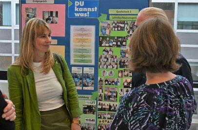 Frau im grünen Blazer im Gespräch mit mehreren Personen vor einer Projektwand des Programms "Du kannst was".
