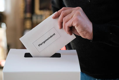 Jemand wirft einen Umschlag mit der Aufschrift "Pfarrgemeinderat" in eine Wahl-Urne