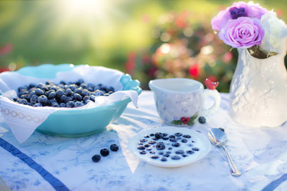 Auf einem Tisch mit weißem Tischtuch, der im Garten zu stehen scheint, eine Schüssel mit Blaubeeren, ein Schälchen mit Blaubeeren in Milch, außerdem ein Kännchen, ein Löffel und eine Vase mit weißen und blassvioletten Blumen.