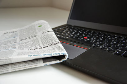 Auf einer weißen Tischoberfläche ein aufgeklappter schwarzer Laptop. Auf der linken vorderen Ecke seiner Tastatur liegt eine zusammengeklappte Zeitung.