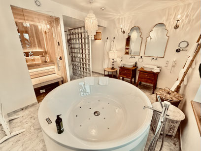 Private Spa Wellness Bad mit großer Sauna und rundem Whirlpool. Luxuriöse Ausstattung mit zwei getrennten Waschbecken, goldenen Spiegeln und Dekoration im Boho Style. Kuschlige Handtücher in ausreichender Zahl und Bademäntel.