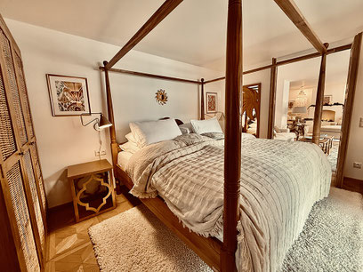 Großes Schlafzimmer mit Himmelbett aus Holz, Flat-TV, großem Kleiderschrank aus Holz. Durch eine weit geöffnete Flügeltür öffnet sich der Blick auf das luxuriöse Wohnzimmer mit dem Kamin.   