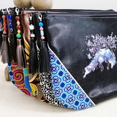 6 grande pochettes plates, en similicuir, pagne/wax et floquées, sont debout appuyées contre une statuette en pierre