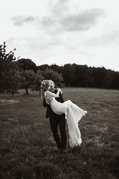 Der Bräutigam trägt seine Braut während des Brautpaarshootings auf dem Arm. Das Brautpaarshooting ist auf einer Wiese bei Erding.