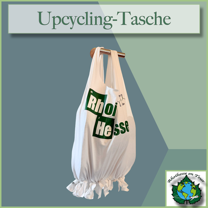 Weiße Upcycling-Tasche aus einem Rhoihesse-T-Shirt, unten mit geknoteten Fransen verschlossen, hängend an einem Holzhalter. Hintergrund in gedeckten Grün- und Blautönen im Rhoihesse-on-Tour-Stil.