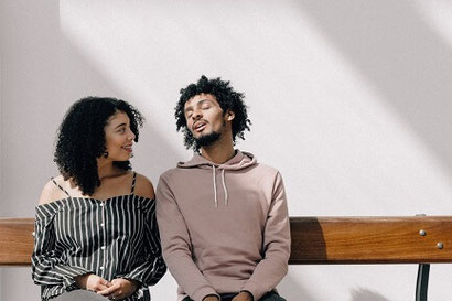 A man and a woman seated on a bench, speaking to each other. The man looks like he may be a little distracted.