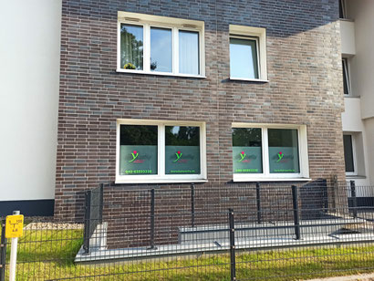 Fassadenansicht eines Gebäudes mit zwei unteren Fenstern, die mit Sichtschutzfolien mit grünem und rotem Logo von Physiofly und Kontaktdaten beklebt sind, davor ein schwarzer Metallzaun