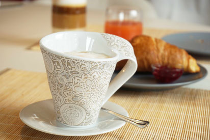 Frühstücksgedeck: Im Vordergrund ist eine verzierte Kaffetasse mit Kaffe zu sehen. Im Mittelgrund ein Croissant mit und im Hintergrund ein Glas Saft und ein Latte macchiato   