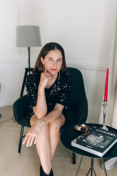 Frau in schwarzem, glitzerndem Kleid sitzt elegant auf einem dunklen Sessel. Sie trägt feinen Silberschmuck, im Vordergrund stehen ein Tisch mit Kerze und einem Chanel-Bildband.