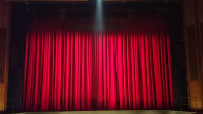 Der im Einlasslicht beleuchtete rote Hauptvorhang des Stadttheaters Bremerhaven. Er scheint rot zu glühen. Von oben kommt ein enger Lichtkegel für den Dirigenten, der später im hier nicht sichtbaren Orchestergraben steht und dirigiert.