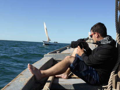 Un homme sur une barque, assis, un appareil photo dans la main. Une autre barque arrive sur la droite de l'image. 