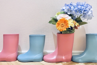 ピンク色と水色のレインブーツが片足ずつ交互に一列に並べられている。ガーベラとアジサイのブーケがレインブーツに飾られている。