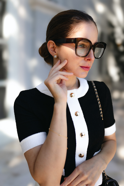 Elegante Frau in schwarz-weißem Outfit mit großen Sonnenbrillen und Perlenohrringen im weichen Tageslicht.