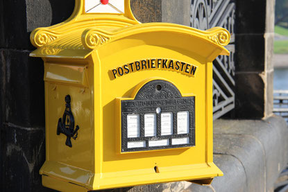 Ein alter gelber Briefkasten aus Metall. Die Oberseite ist leicht gebogen und hat Schnörkel. Auf der Vorderseite steht in Schwarz "Postbriefkasten", darunter sind Felder für die Leerungszeiten. An der linken Wand ist das Symbol eines Posthorns zu sehen.