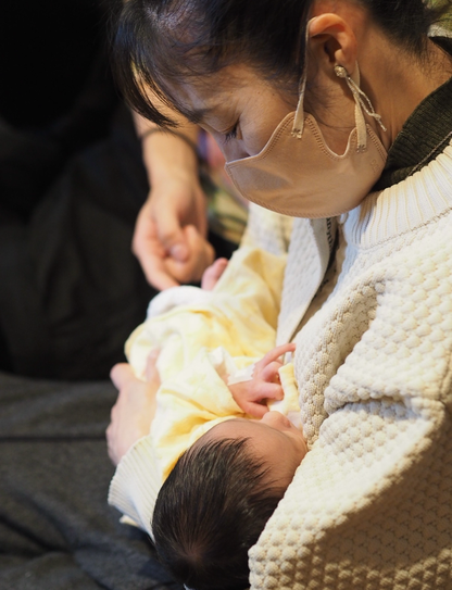髪を一つに束ねマスクをした女性の腕には生まれたばかりの赤ちゃんを抱き、女性の耳にはクラゲがモチーフのビーズアクセサリーがついています。初孫を抱く彼女はとても嬉しそうな笑顔です。