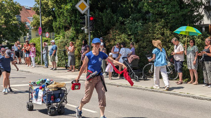 Stefanie (eine Frau mit VfR T-Shirt) zieht einen mit vielen Wasserflaschen gefüllten Bollerwagen durch eine Straße und schwenkt gleichzeitig noch ein Theraband. An der Seite stehen Zuschauer und auf dem Wagen steht "Löschfahrzeug".