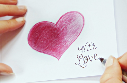 Auf ein weißes Blatt hat eine Hand gerade "With Love" geschrieben. Eine 2. Hand hält das Papier fest. Über der Schrift ein mit Wachskreide handgemaltes Herz mit Farbverlauf von Beere nach Rosa.