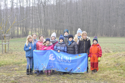 Eine Kindergruppe steht auf einer Wiese vor einem Waldstück. Sie halten dabei einen blauen NAJU Banner hoch.