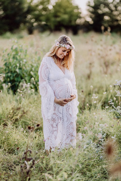 Blonde Frau in weissem Kleid aus Spitze schaut lächelnd auf ihren schwangeren Bauch