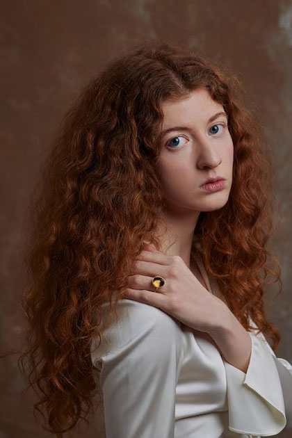 Frau mit langen roten Locken trägt einen goldenen MOKOŠEV Ring mit dunklem Stein vor warmem, neutralem Hintergrund.